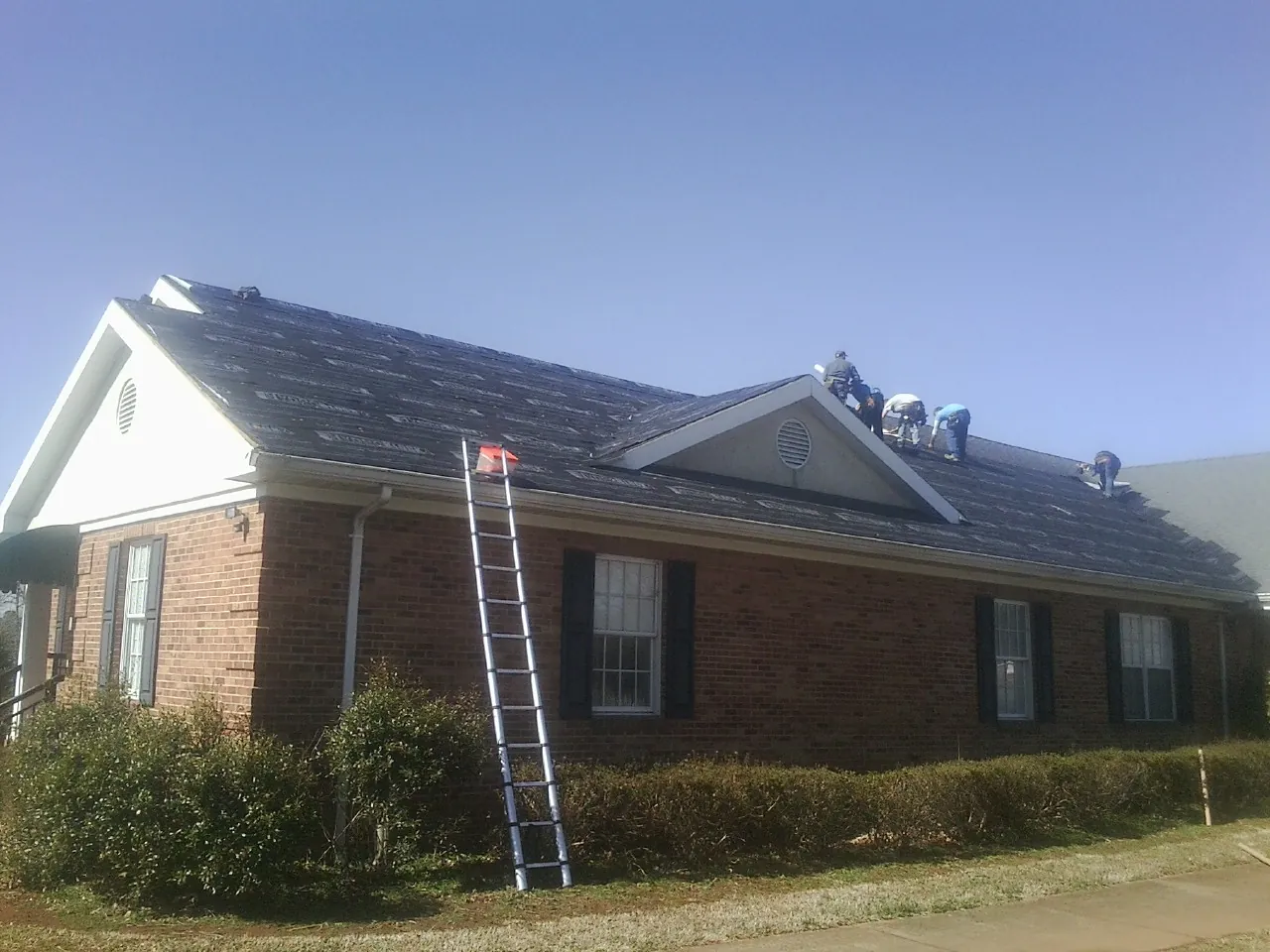 Crew tearing shingles off a brick ranch roof with a ladder set — Metal Roof Repair work by our crew in Luck