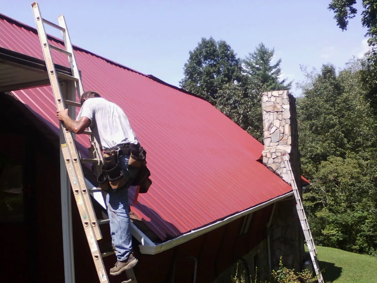 Roofer on a ladder beside a red metal roof and stone chimney — Soffit & Fascia Repair by Roofing Brothers in Luck